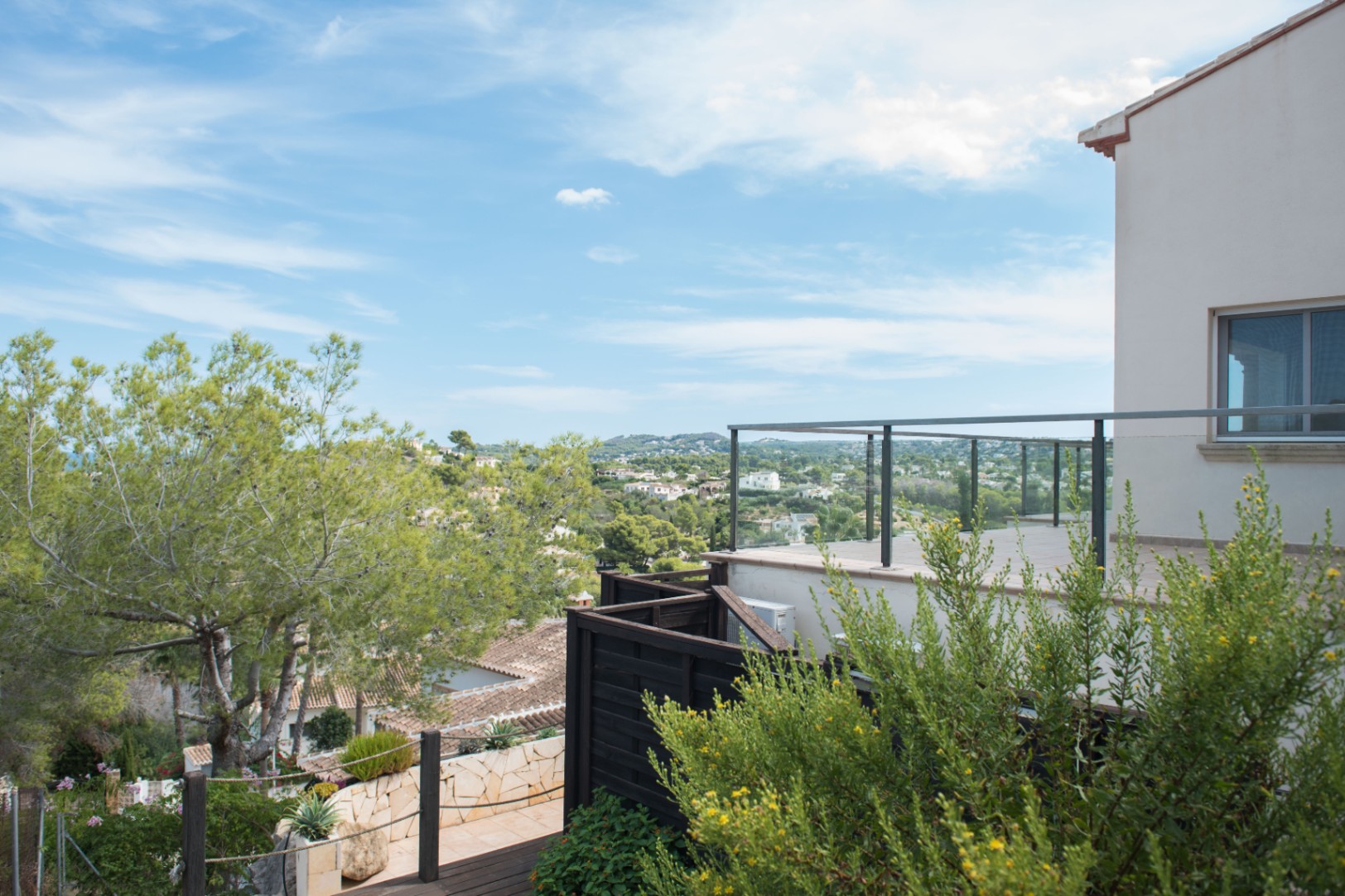 Elevated terrace with glass railings offering panoramic views of the surrounding valley and Mediterranean vegetation under a clear sky.