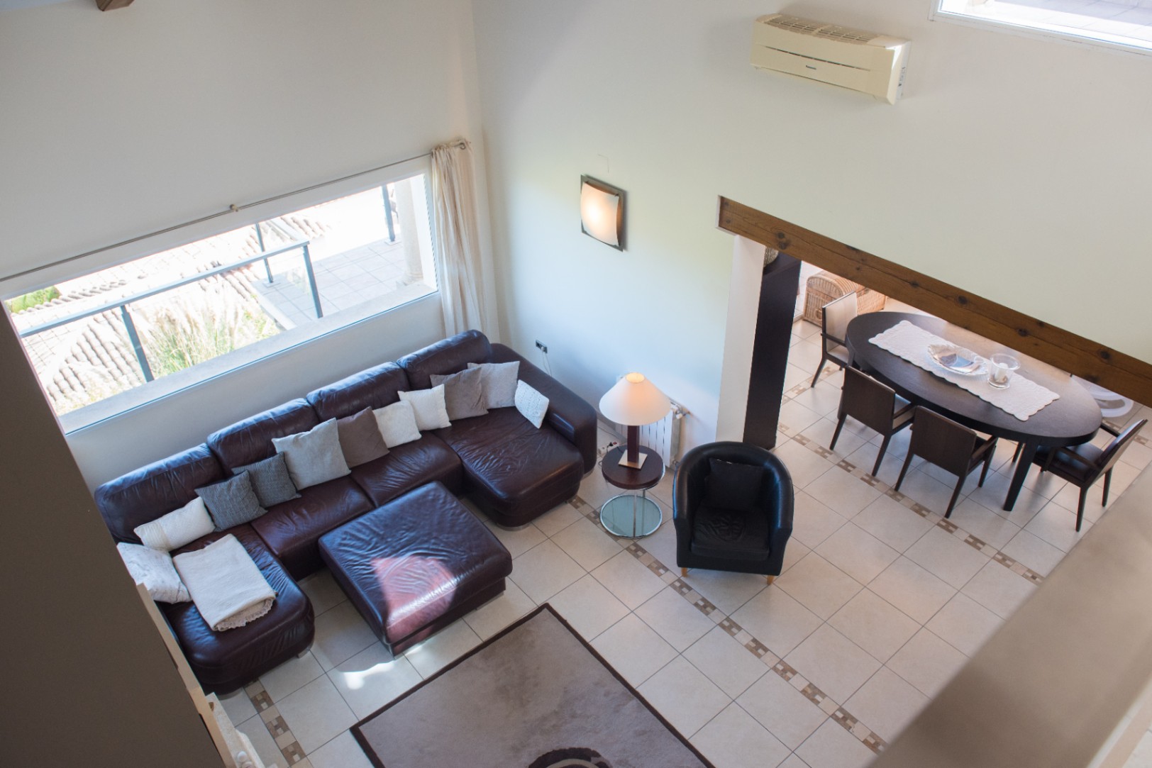 Elevated view of a living area featuring a large brown leather sectional, tiled flooring, and an adjacent dining space with a dark wood table under a timber beam.