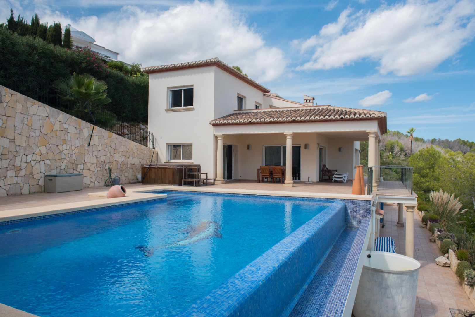 Exterior view of a white villa featuring an infinity-style pool with blue mosaic tiles and a dolphin motif, adjacent to a covered terrace with columns.