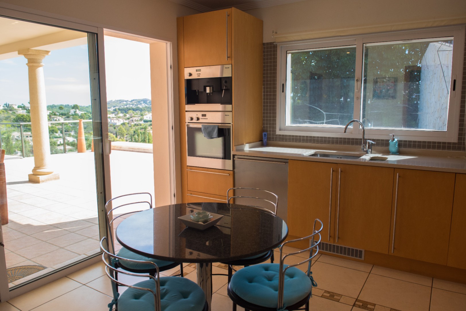 Kitchen with light wood cabinetry, integrated Miele appliances, and a granite-top dining table. A sliding glass door opens to a terrace with panoramic valley views.