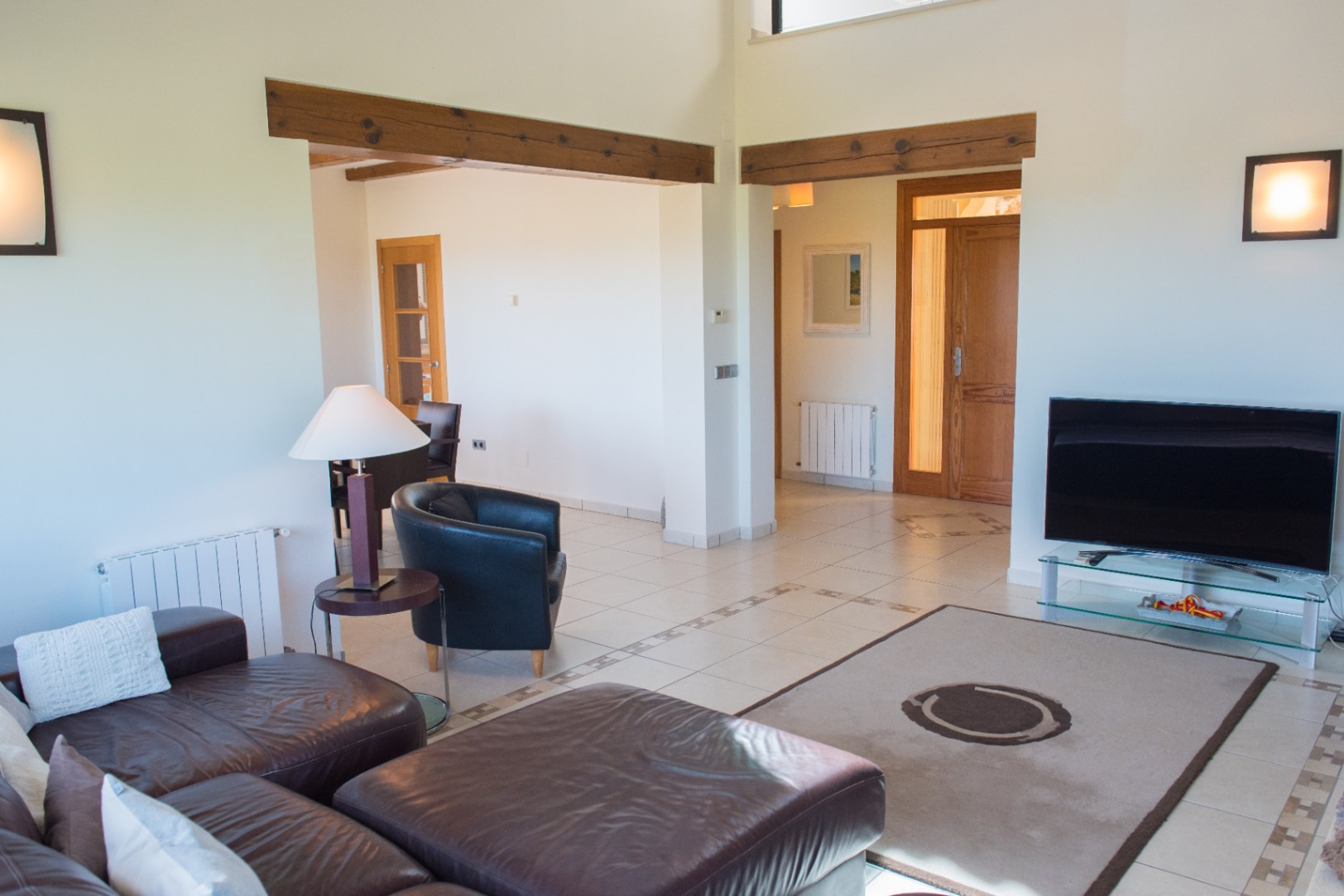 Living room featuring dark leather sofas, tiled floors with decorative inlays, and exposed wooden beams. The open layout leads to a dining area and wooden interior doors.