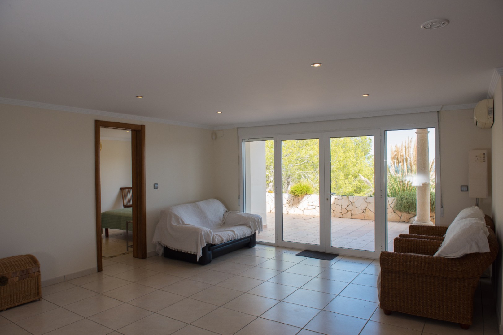 Living room with tiled floors and large glass doors opening to a terrace. Features wicker armchairs, recessed lighting, and views of a Mediterranean garden.