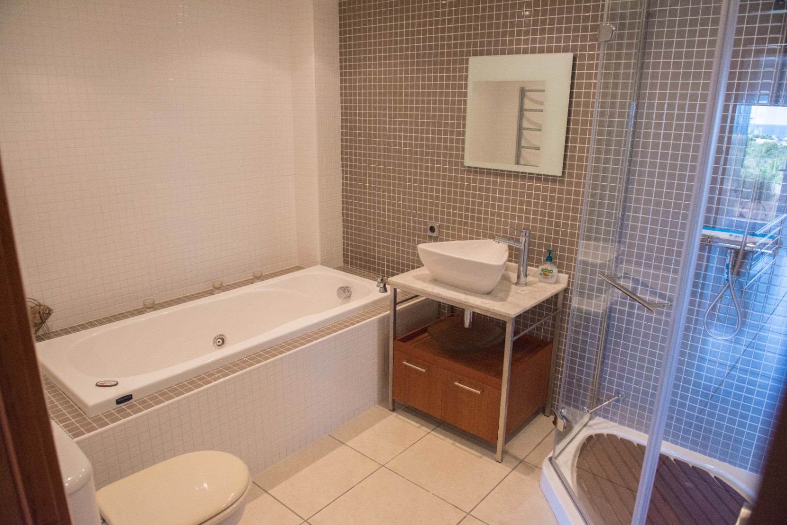 Modern bathroom featuring a built-in bathtub, a glass-enclosed shower with wooden flooring, and a vessel sink on a marble-topped vanity with taupe mosaic tiles.