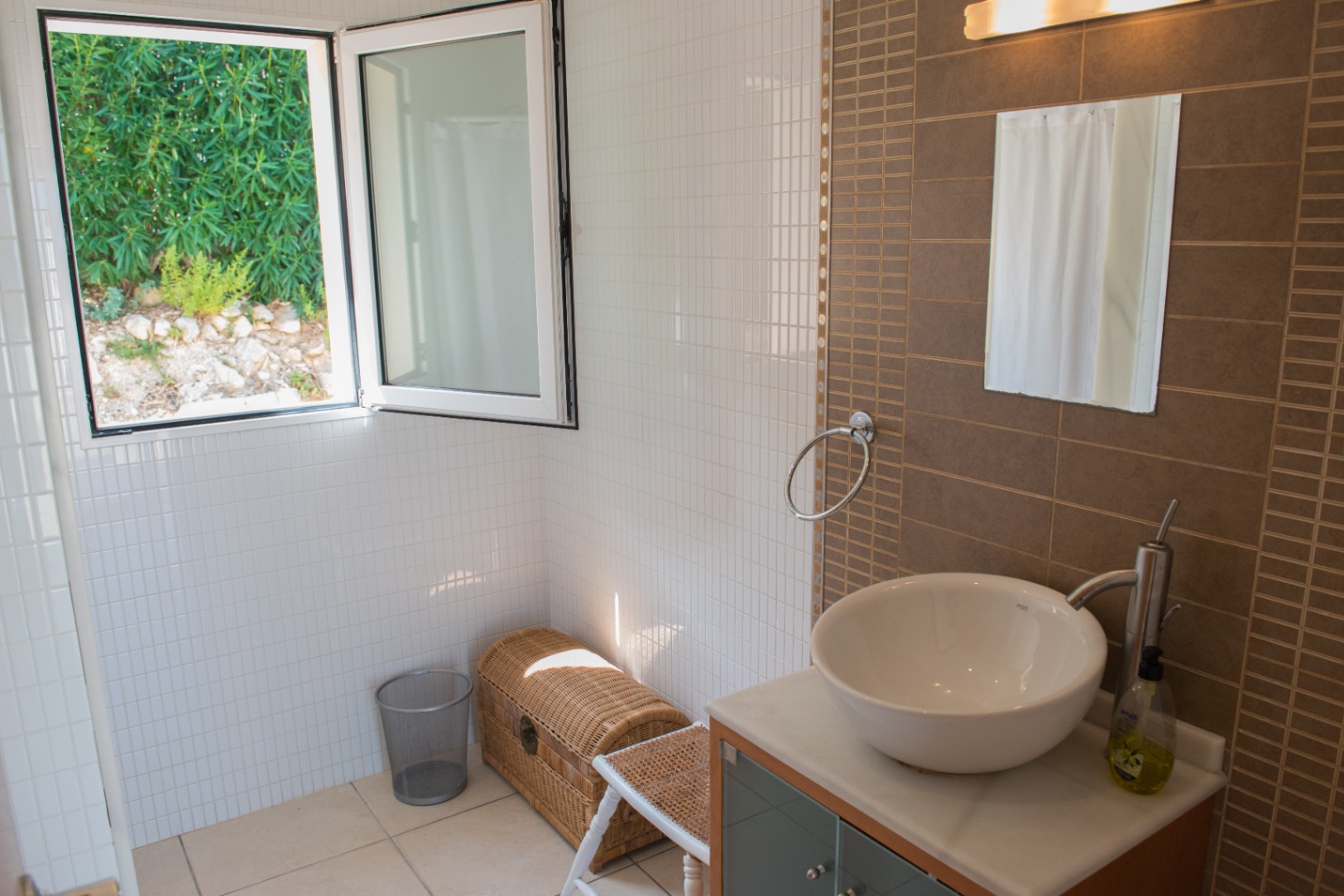 Modern bathroom featuring a white vessel sink, brown tiled accent wall, and an open window providing natural light and views of the garden foliage.
