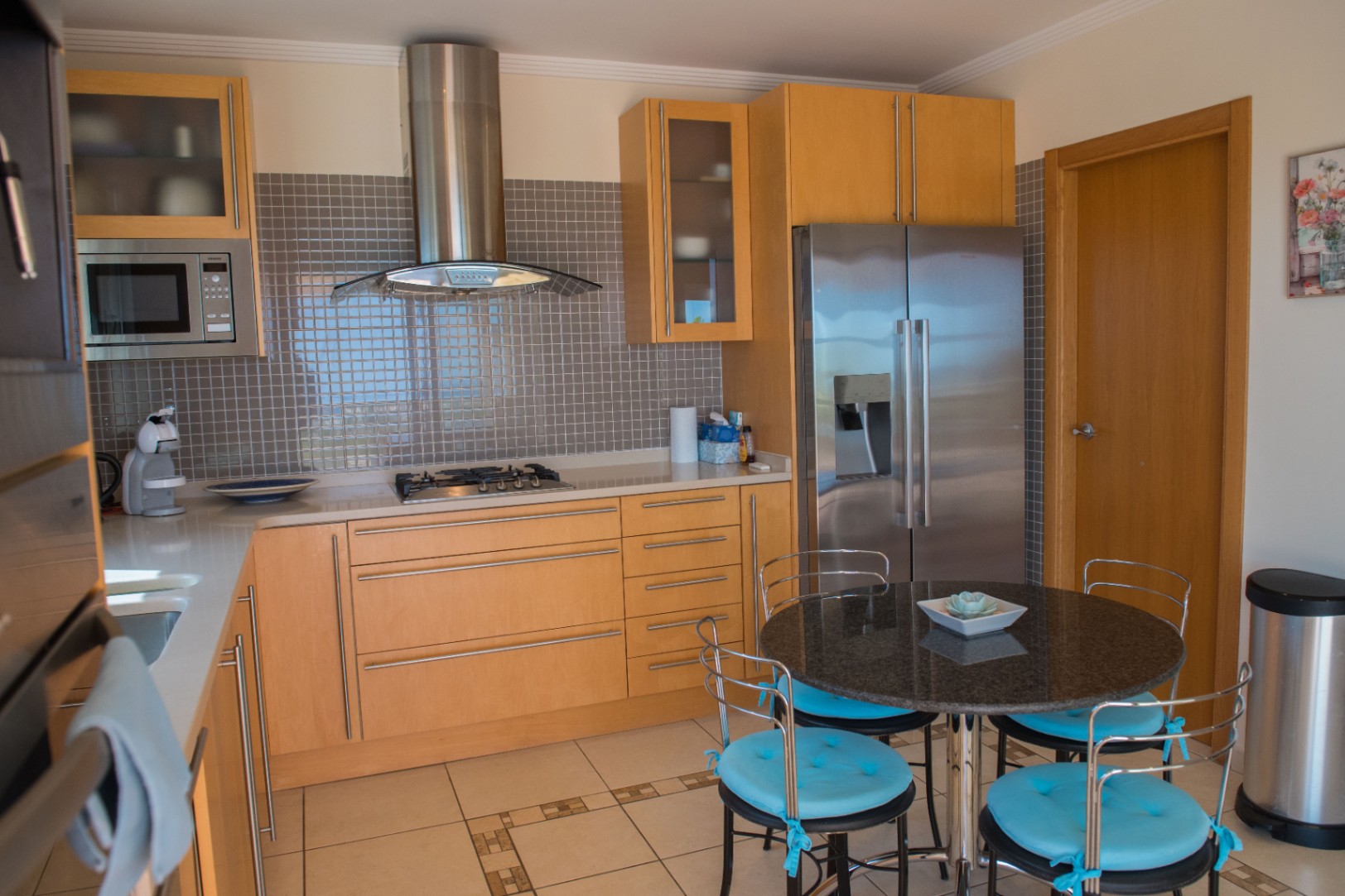 Modern kitchen featuring light wood cabinetry, a grey mosaic backsplash, and stainless steel appliances. Includes a round granite dining table with blue-cushioned chairs.