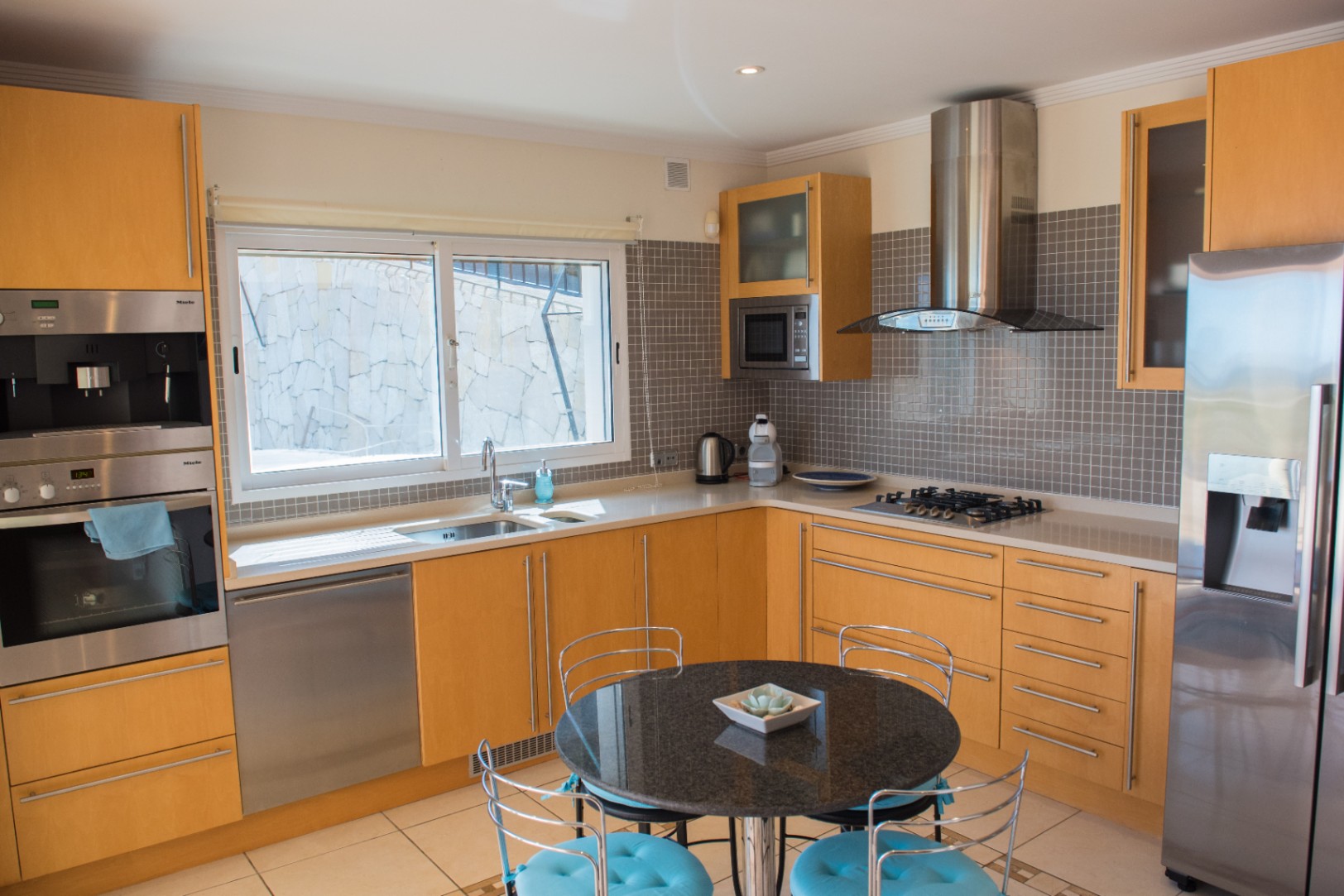 Modern kitchen featuring light wood cabinetry, stainless steel Miele appliances, and a grey mosaic backsplash. Includes a black granite breakfast table and gas cooktop.
