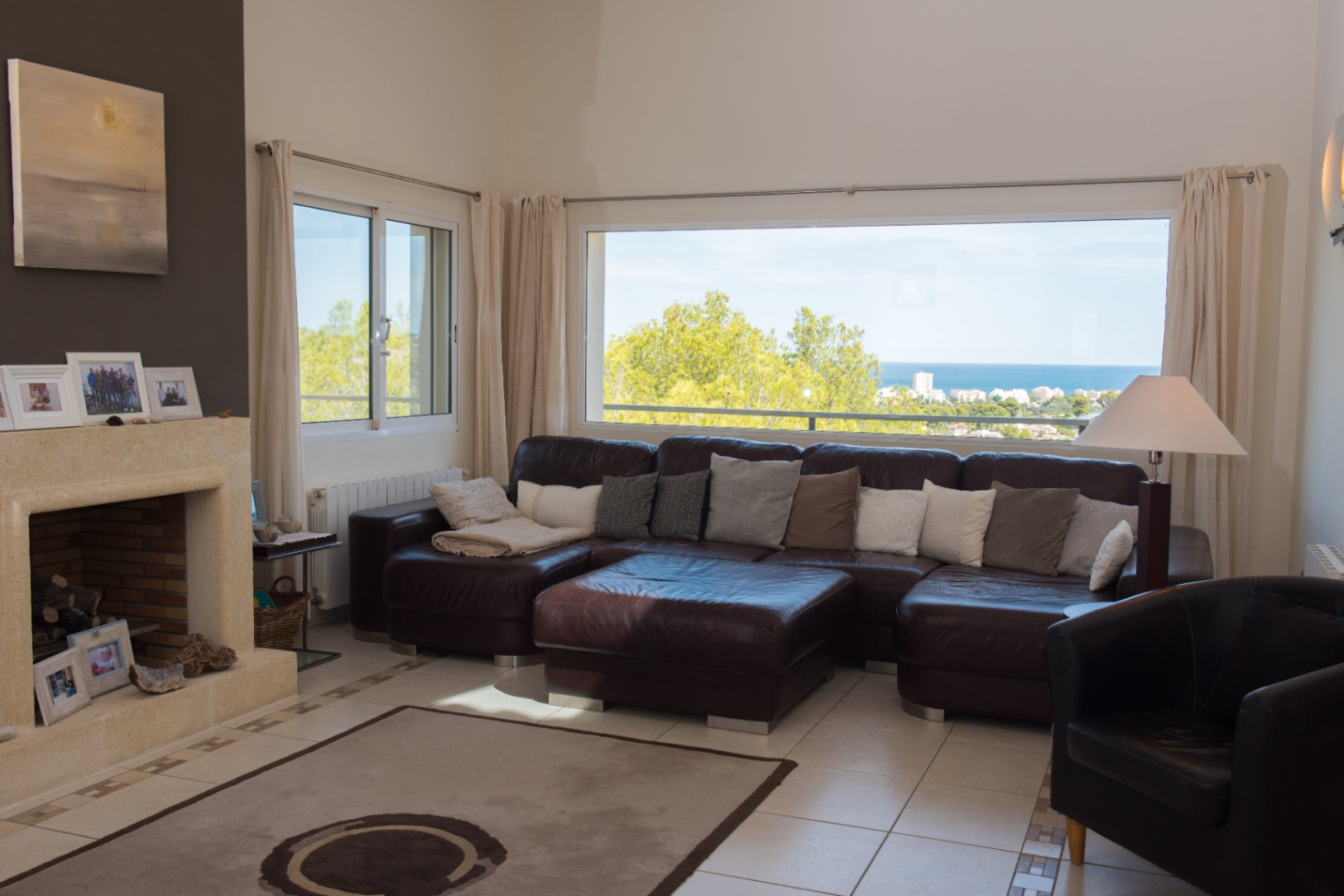 Spacious living room featuring a large brown leather sectional, a stone fireplace, and a wide panoramic window with views of the coast and sea.