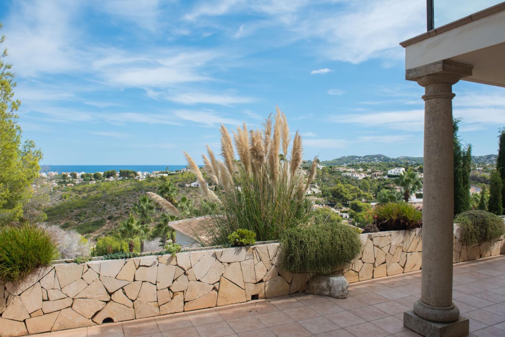 Terrace with stone columns and a dry-stone perimeter wall, featuring pampas grass and panoramic views of the Mediterranean coastline and surrounding hills.
