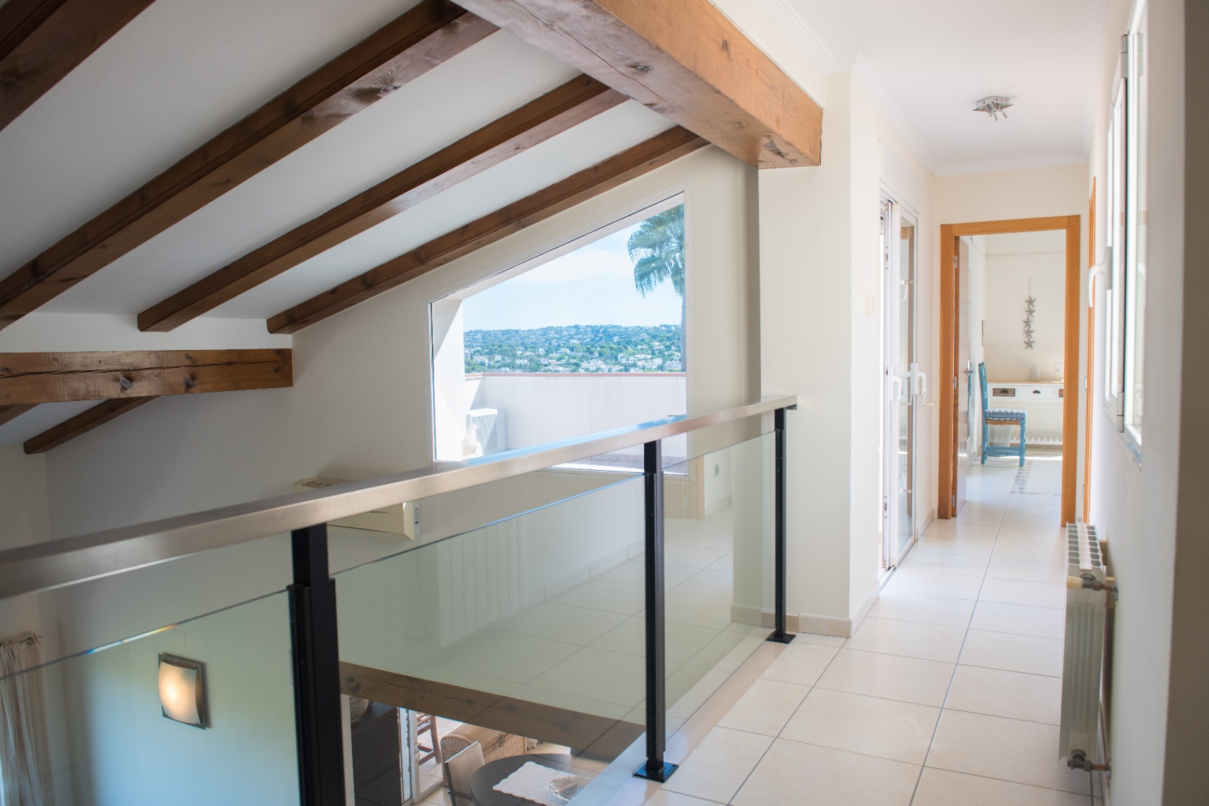 Upper-level landing featuring exposed dark wood ceiling beams, a glass balustrade, and a large trapezoidal window with views of the surrounding landscape.