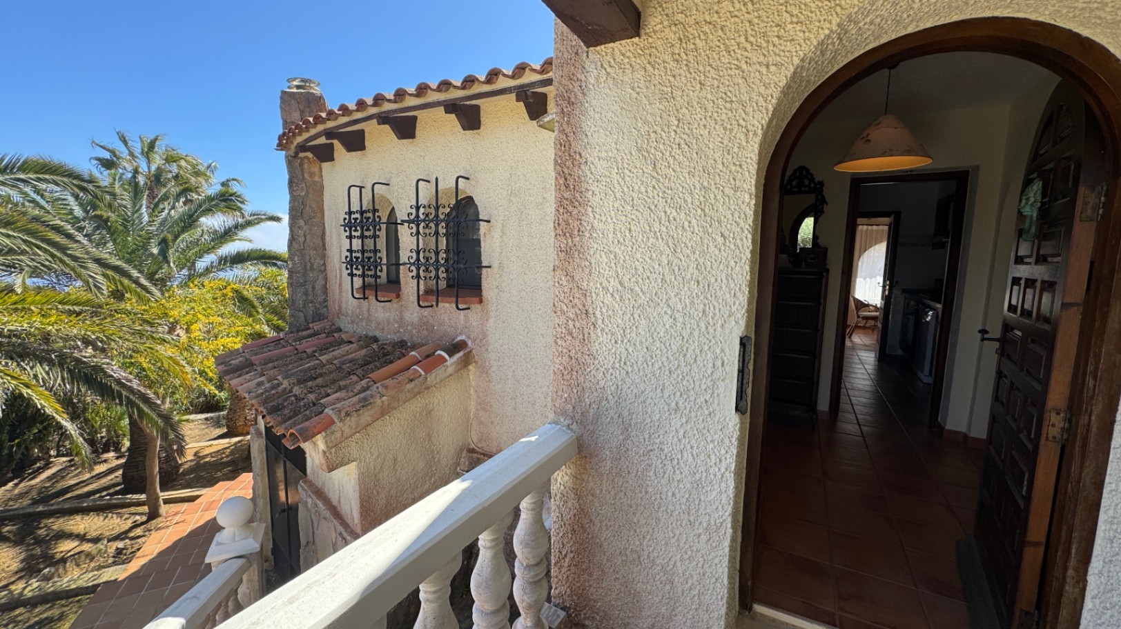 Arched villa entrance featuring a solid wood door, terracotta tiling, and decorative wrought iron window grilles set against Mediterranean palms.