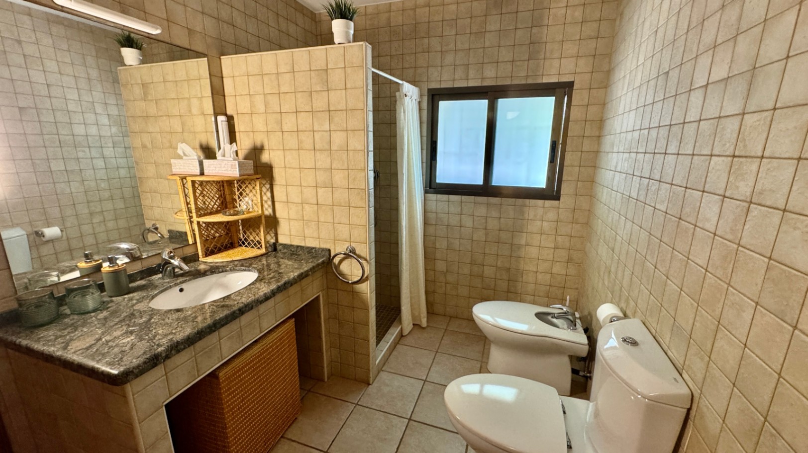 Bathroom featuring a granite vanity with integrated sink and wicker storage. Includes beige wall tiles, a walk-in shower with curtain, and a toilet with bidet.