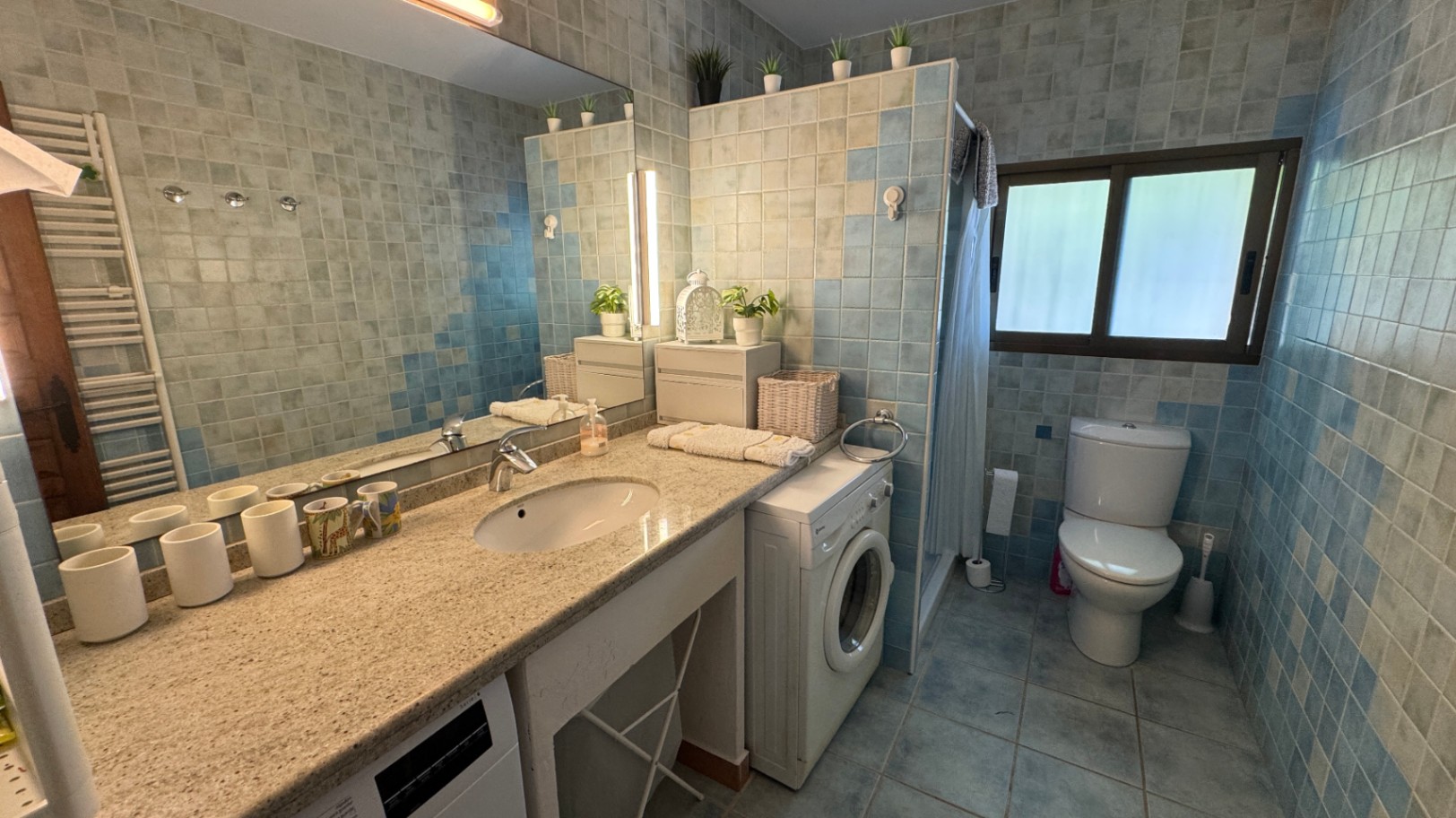 Bathroom with a granite vanity, integrated sink, blue mosaic tiling, and a washing machine. Includes a large mirror and natural light from a side window.