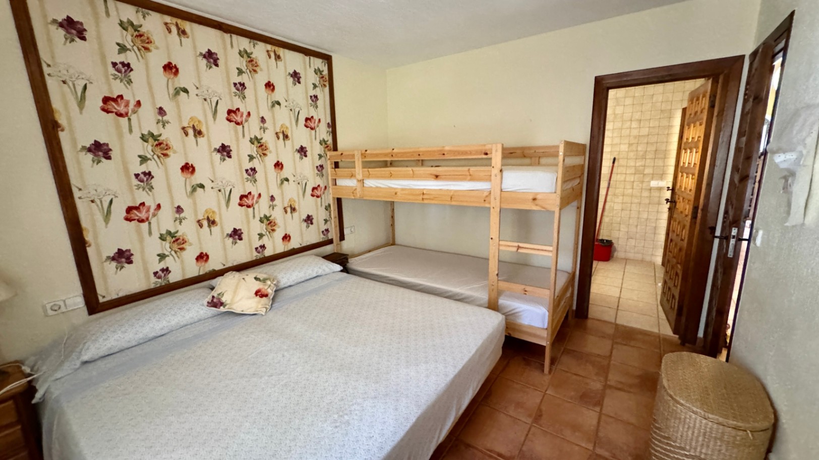 Bedroom featuring a double bed and wooden bunk beds, accented by a floral wallpaper panel and terracotta tile flooring. An ensuite bathroom is visible.