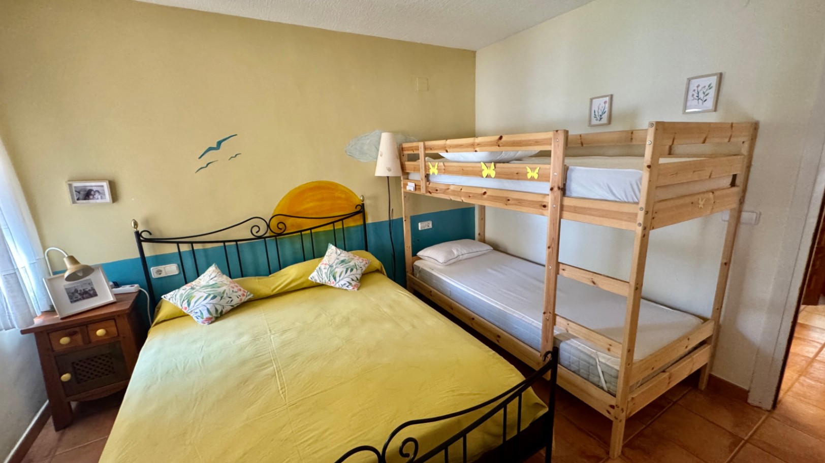 Bedroom featuring a double bed with a black wrought iron headboard and a light wood bunk bed. The room includes terracotta tile flooring and a decorative wall mural.
