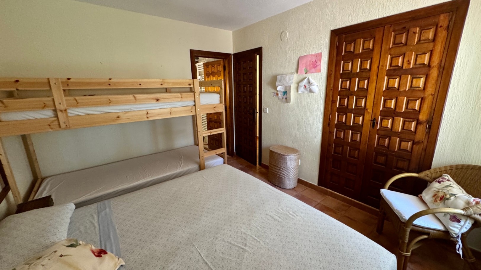 Bedroom featuring pine bunk beds, a built-in wooden wardrobe with paneled doors, and terracotta tile flooring. Includes a wicker armchair and textured walls.