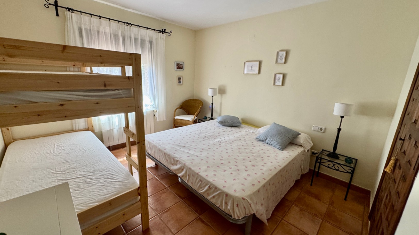 Bedroom featuring wooden bunk beds and a double bed, terracotta tile flooring, and natural light. Includes a wicker armchair and minimalist wall decor.
