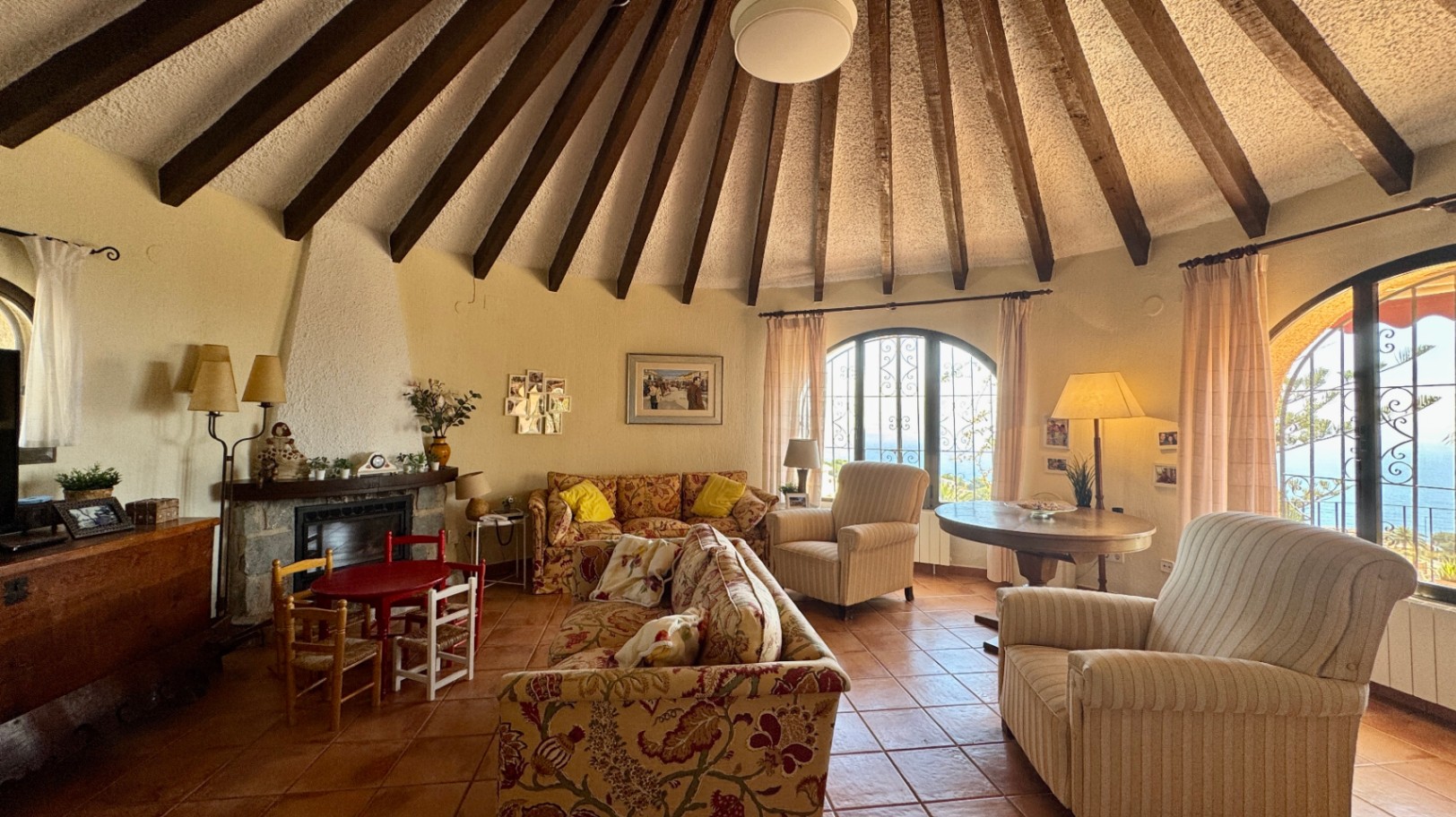 Circular living room featuring a vaulted ceiling with radial wooden beams, terracotta flooring, a stone fireplace, and arched windows overlooking the sea.