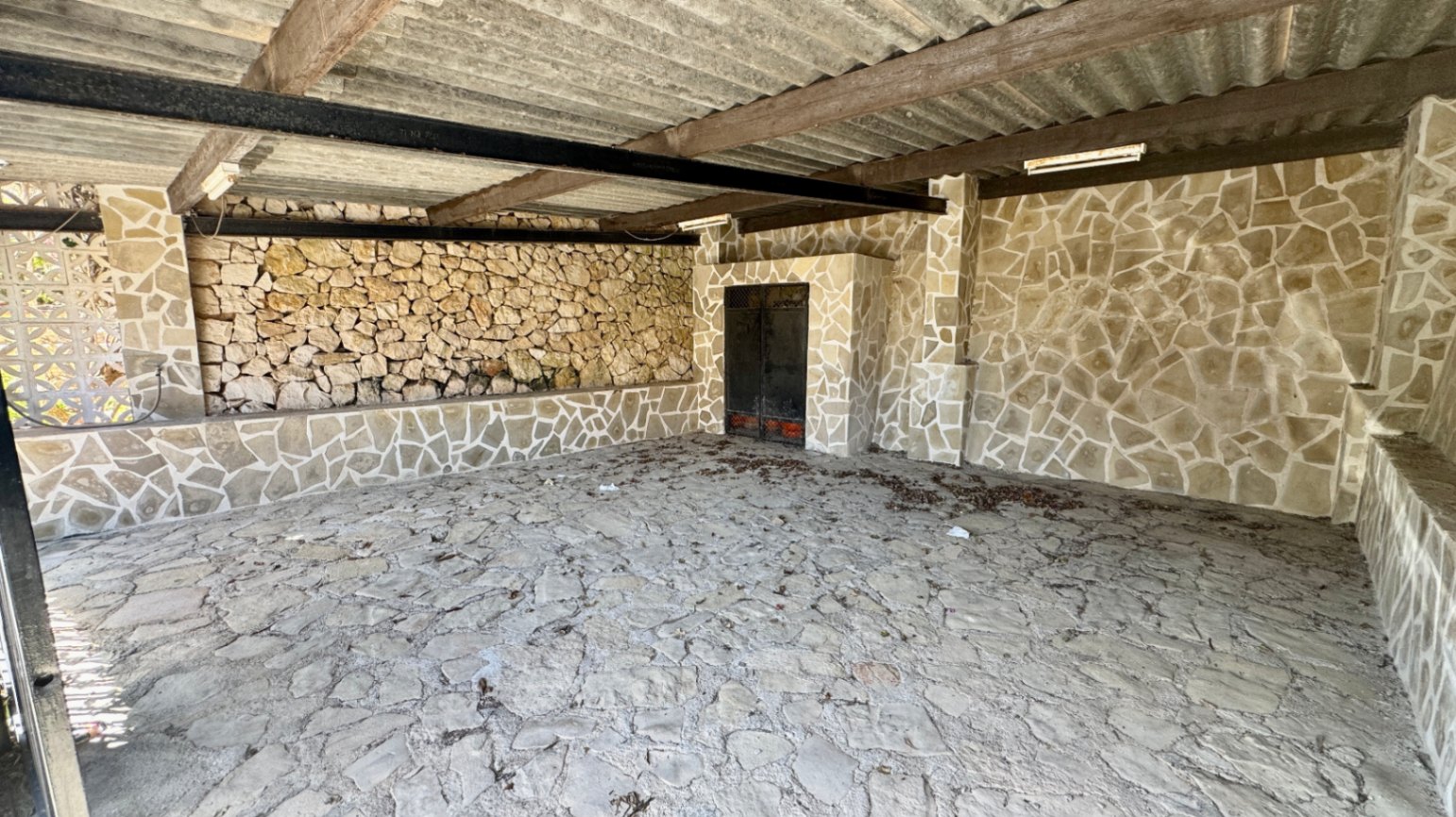 Covered outdoor space featuring rustic stone-clad walls and flooring, supported by wooden and metal beams under a corrugated roof.