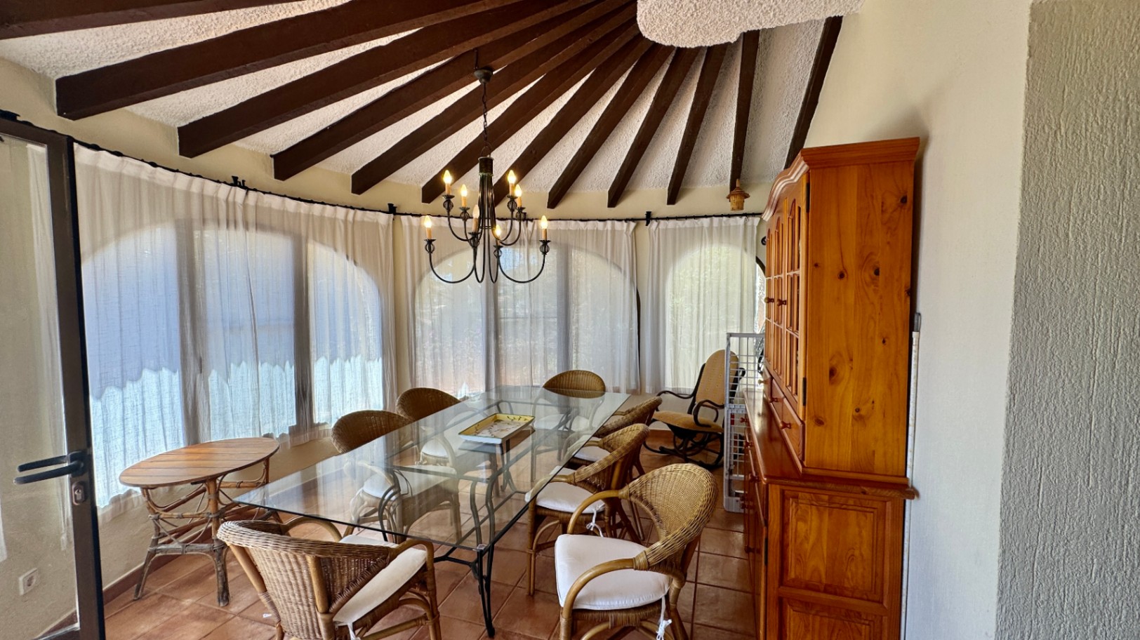 Dining area featuring a radial dark wood beam ceiling, a glass-top table with wicker seating, and a traditional wooden hutch against terracotta flooring.