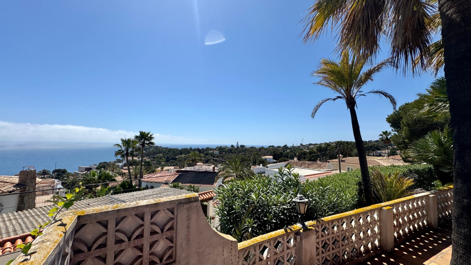 Elevated terrace view overlooking a Mediterranean residential area with terracotta roofs, palm trees, and a distant sea horizon under a clear sky.