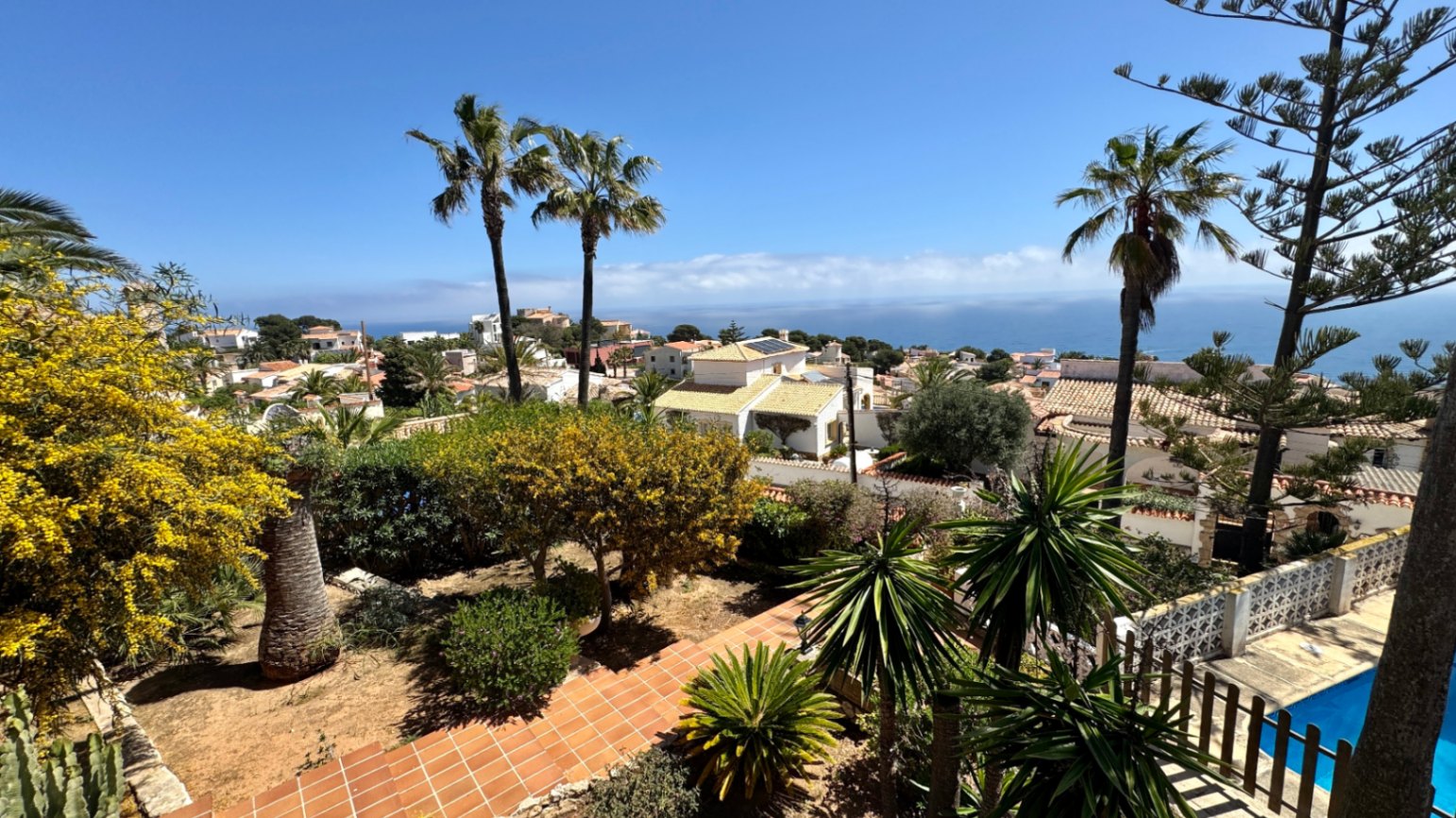 Elevated view of a Mediterranean garden with terracotta paths and palm trees, overlooking a coastal residential area and the open sea.