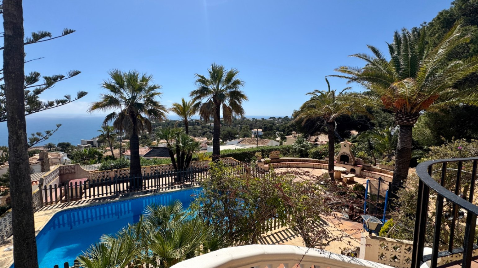 Elevated view of a private swimming pool and stone patio featuring an outdoor fireplace, surrounded by palm trees with distant Mediterranean sea views.