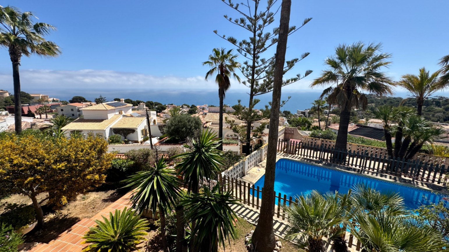 Elevated view of a private swimming pool with a wooden safety fence, surrounded by palm trees and Mediterranean gardens overlooking the sea.