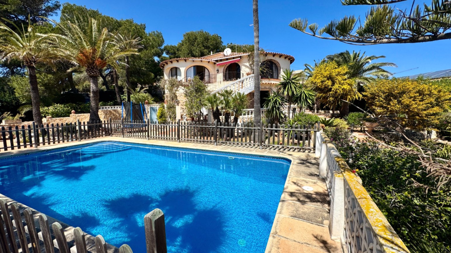 Exterior view featuring a rectangular swimming pool with wooden fencing, set against a Mediterranean villa with arched balconies and palm trees.