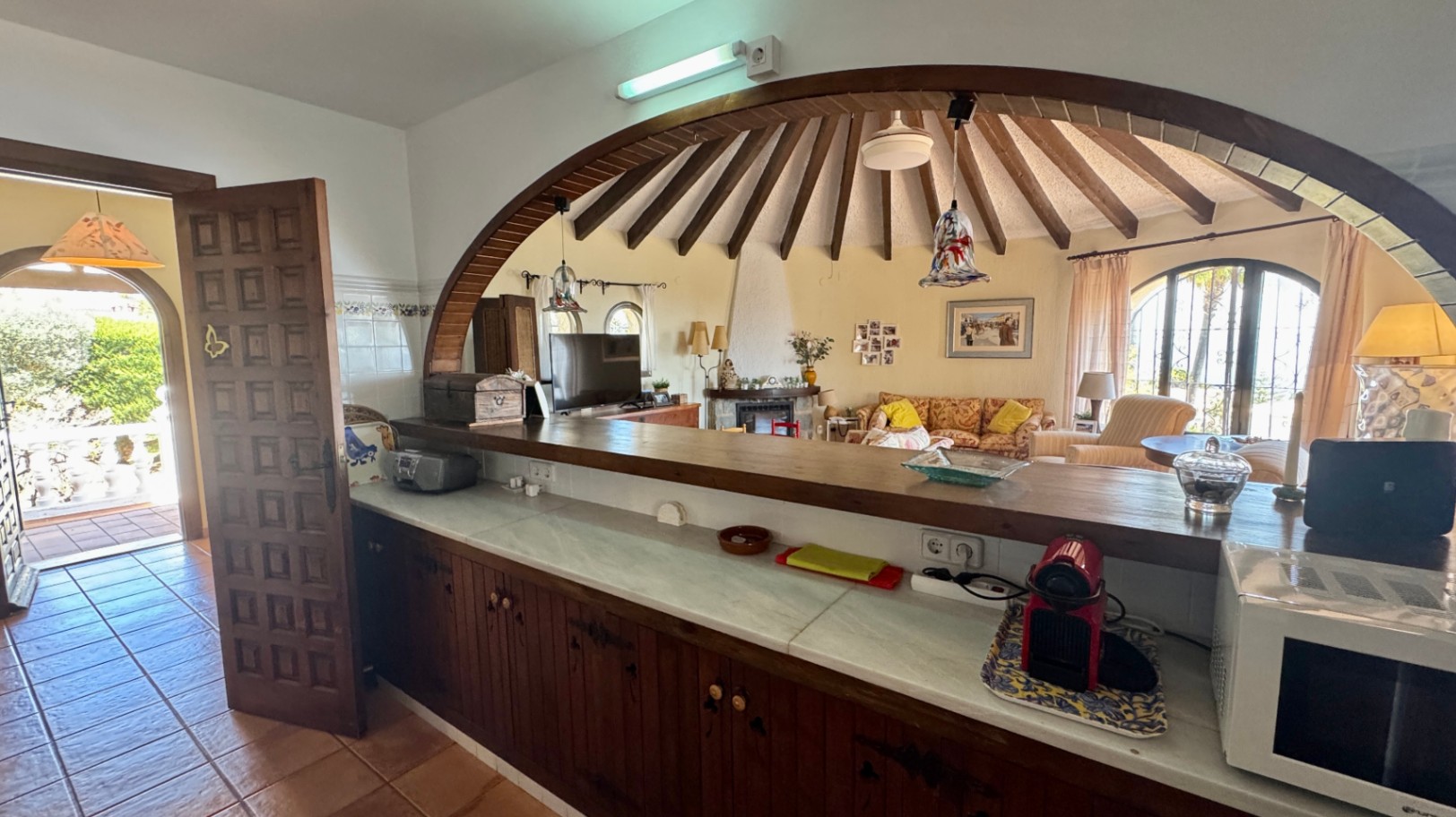 Kitchen bar with marble countertop and dark wood cabinets, looking through a large wooden arch into a circular living room with radial ceiling beams.