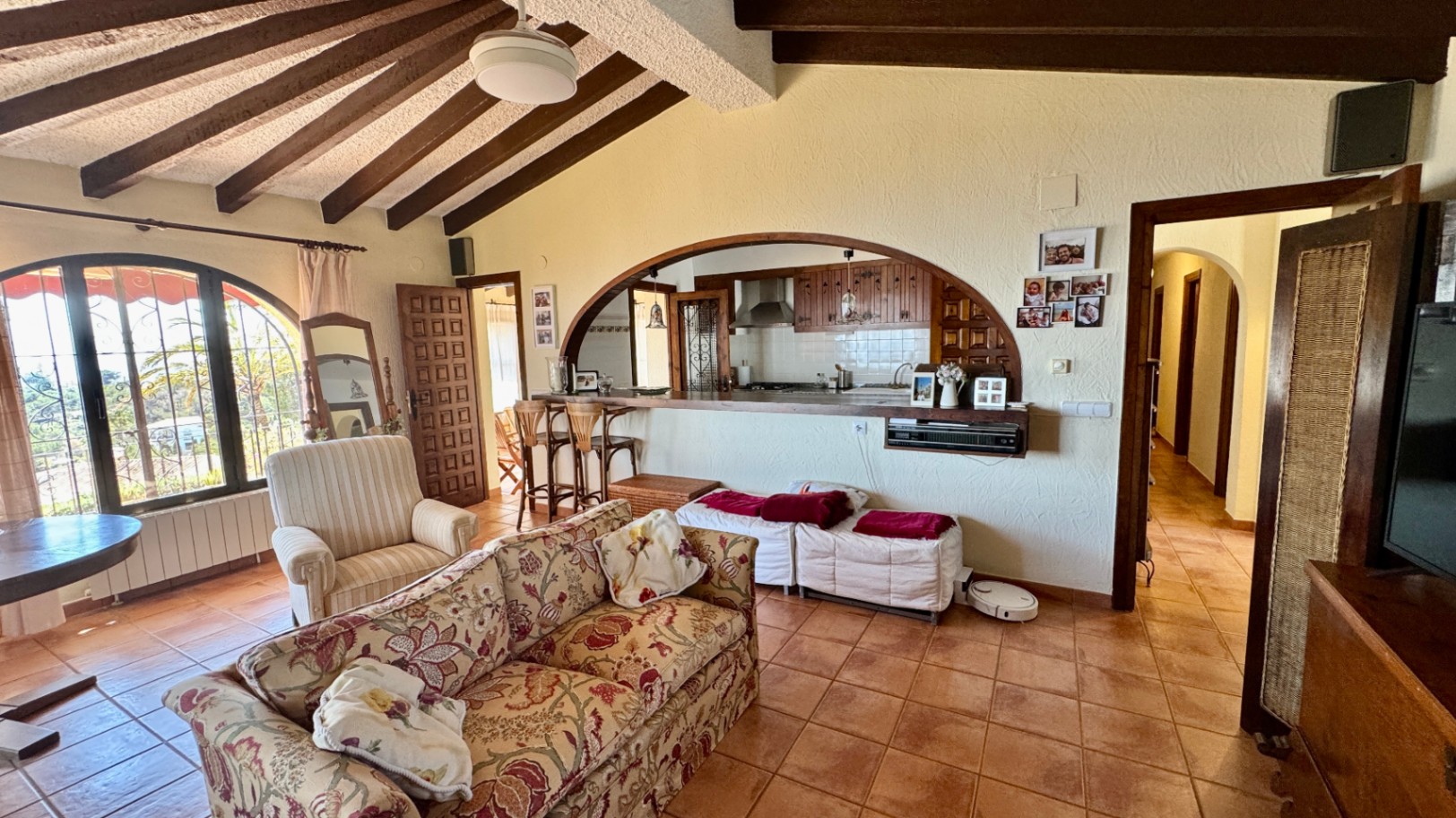 Living area featuring sloped ceilings with dark wood beams, terracotta flooring, and a wide arched opening connecting to the kitchen with a wooden breakfast bar.