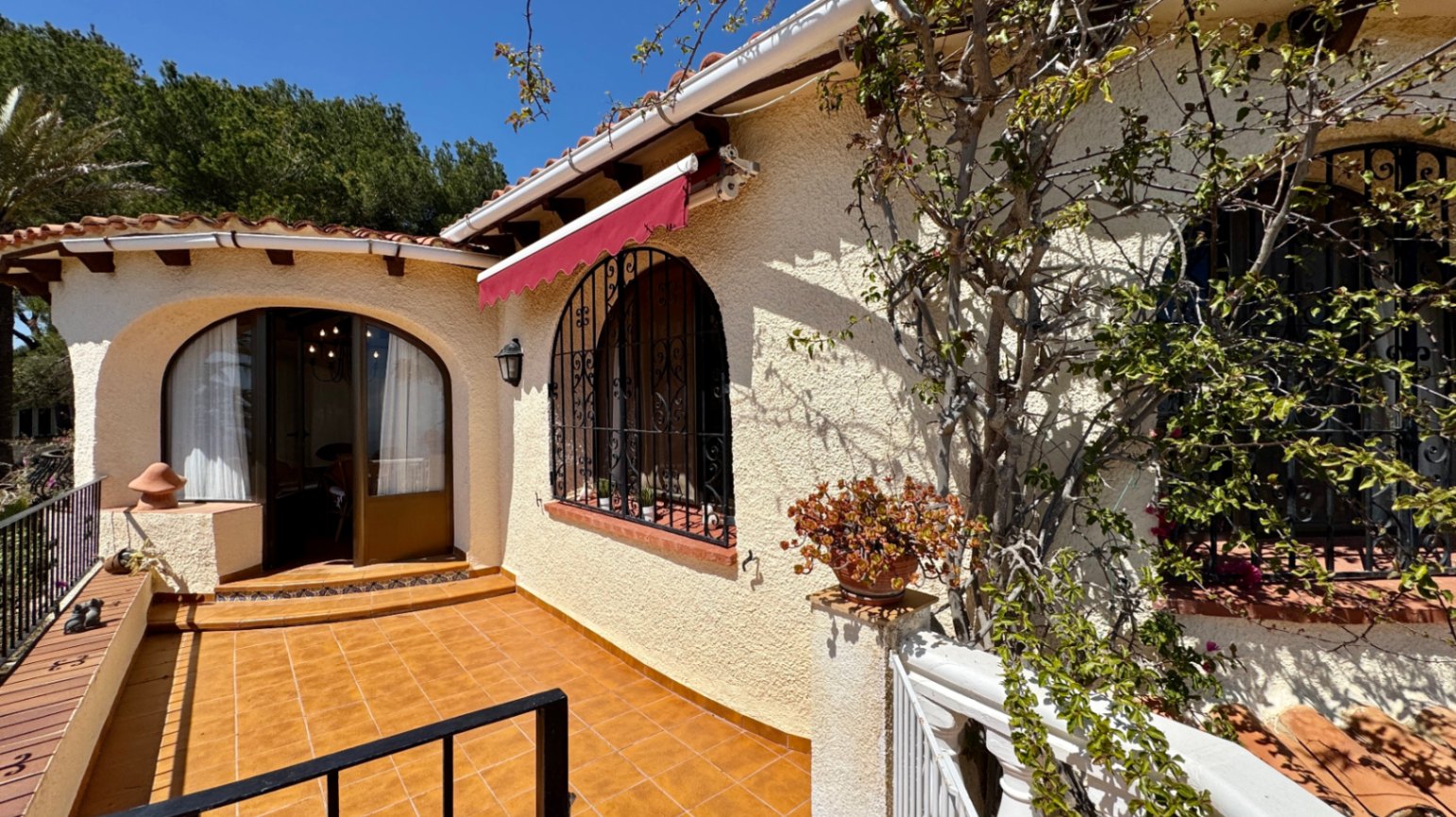 Mediterranean villa exterior with a terracotta tiled terrace, arched windows featuring wrought iron grilles, and a red retractable awning.