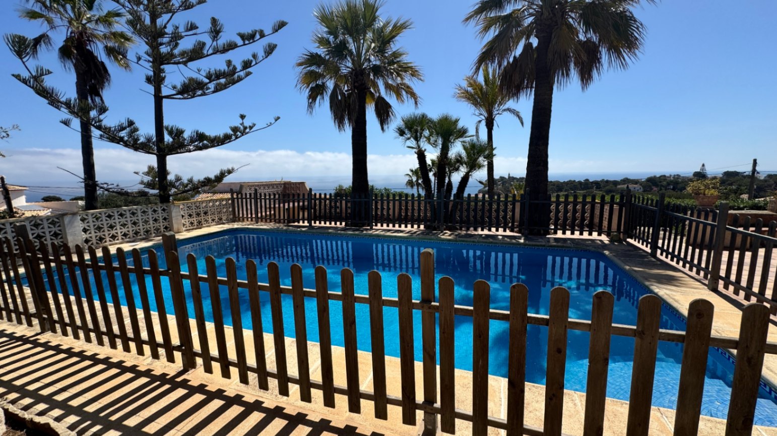 Outdoor swimming pool with blue tiling and a wooden safety fence. The area features mature palm trees and elevated views of the Mediterranean coastline.