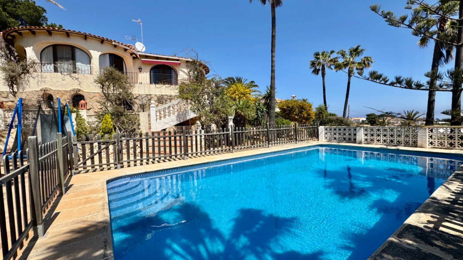 Outdoor swimming pool with blue tiling and integrated steps, surrounded by a wooden fence and a Mediterranean villa featuring stone cladding and arched balconies.
