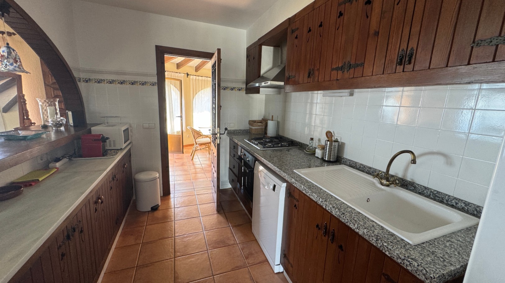 Rustic galley kitchen featuring dark wood cabinetry, granite countertops, and terracotta tile flooring. Includes a gas cooktop and a pass-through archway.