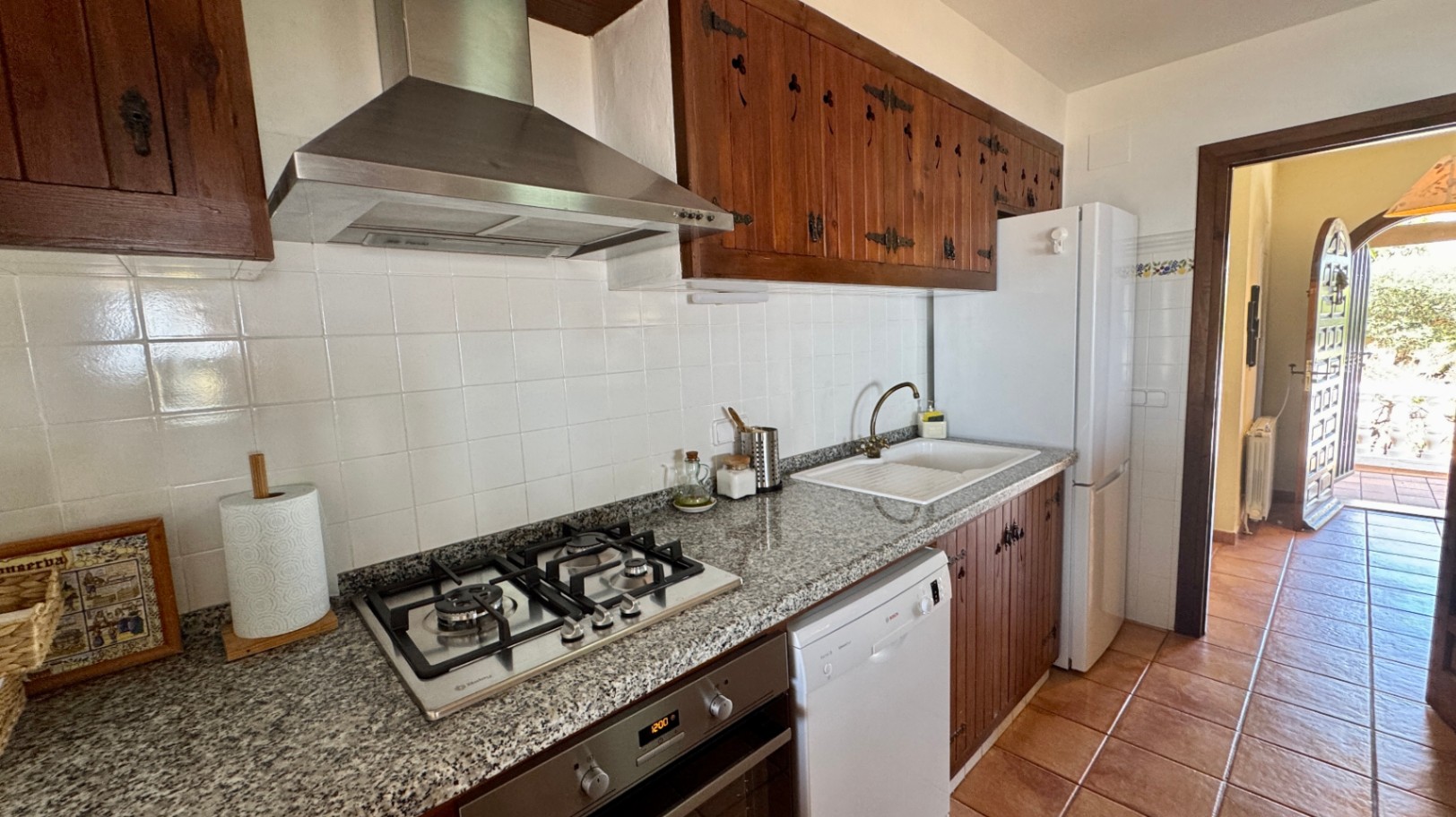 Rustic kitchen featuring dark wood cabinetry with ornate hinges, a granite countertop, gas stovetop, and white tiled walls. The space includes a white dishwasher and terracotta flooring.