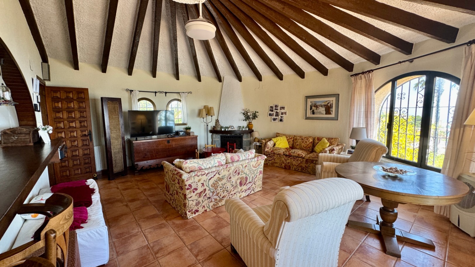 Spacious living room featuring a vaulted ceiling with radial wooden beams, terracotta tile flooring, a fireplace, and arched windows with decorative ironwork.
