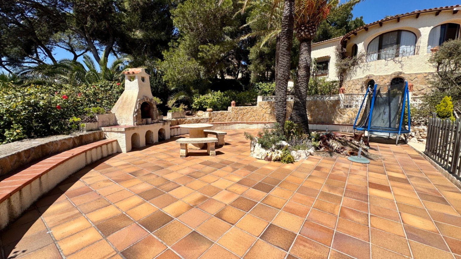 Spacious terracotta terrace featuring a built-in stone barbecue, stone seating area, and palm trees against a Mediterranean villa backdrop.