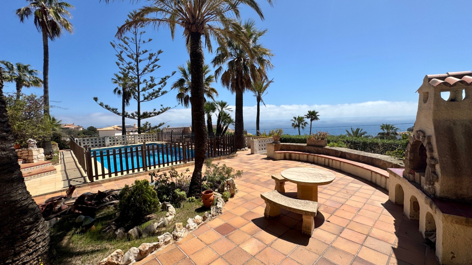 Terracotta tiled terrace featuring a stone dining set, a rustic outdoor fireplace, and a fenced swimming pool overlooking the sea and palm trees.