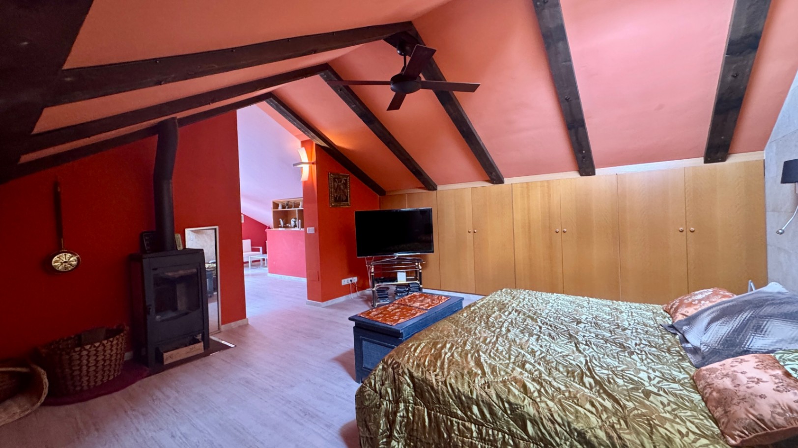 Attic bedroom featuring exposed dark wood beams, terracotta-toned walls, a wood-burning stove, and integrated light wood wardrobes.