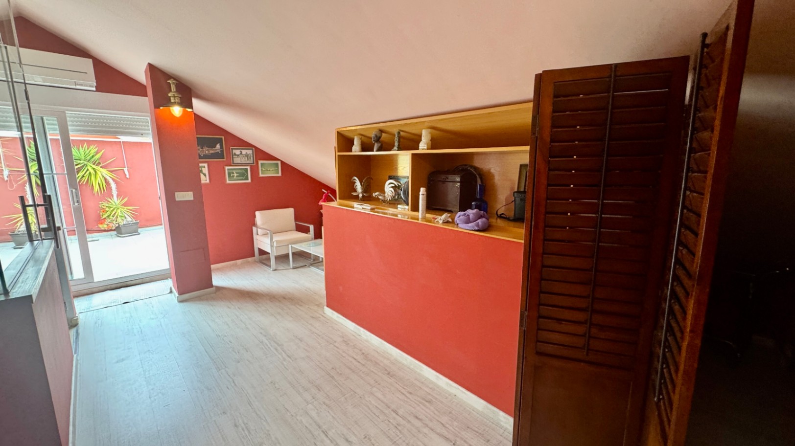Attic living area with terracotta walls, sloped ceilings, and built-in wooden shelving. A glass door opens to a private terrace with Mediterranean plants.