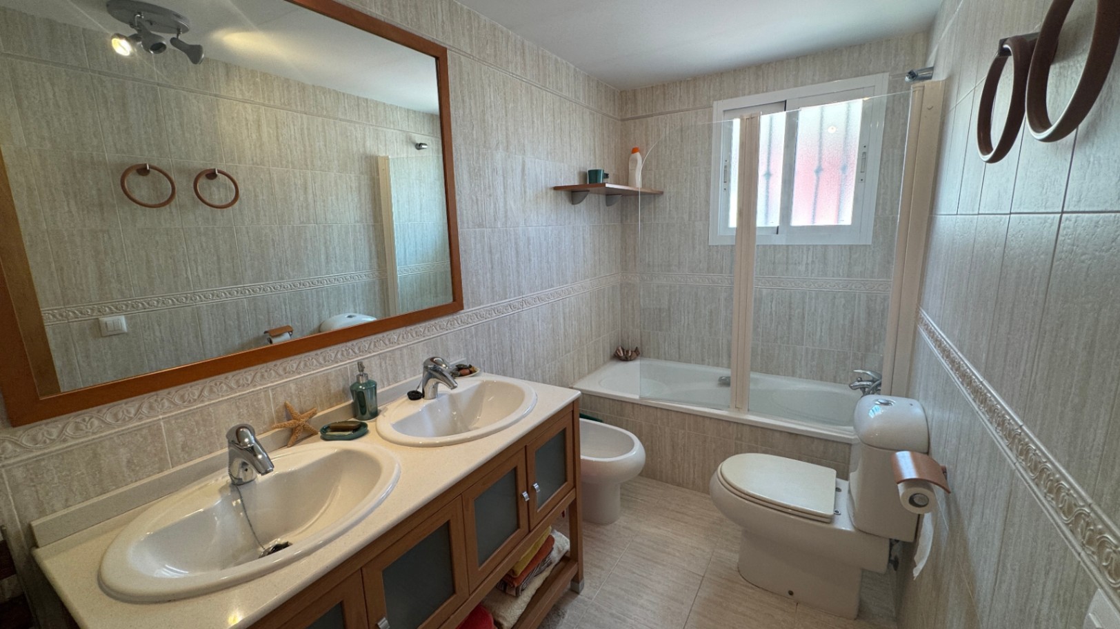 Bathroom featuring a double vanity with wooden cabinetry, a large framed mirror, and a bathtub with a glass partition and neutral tiled walls.
