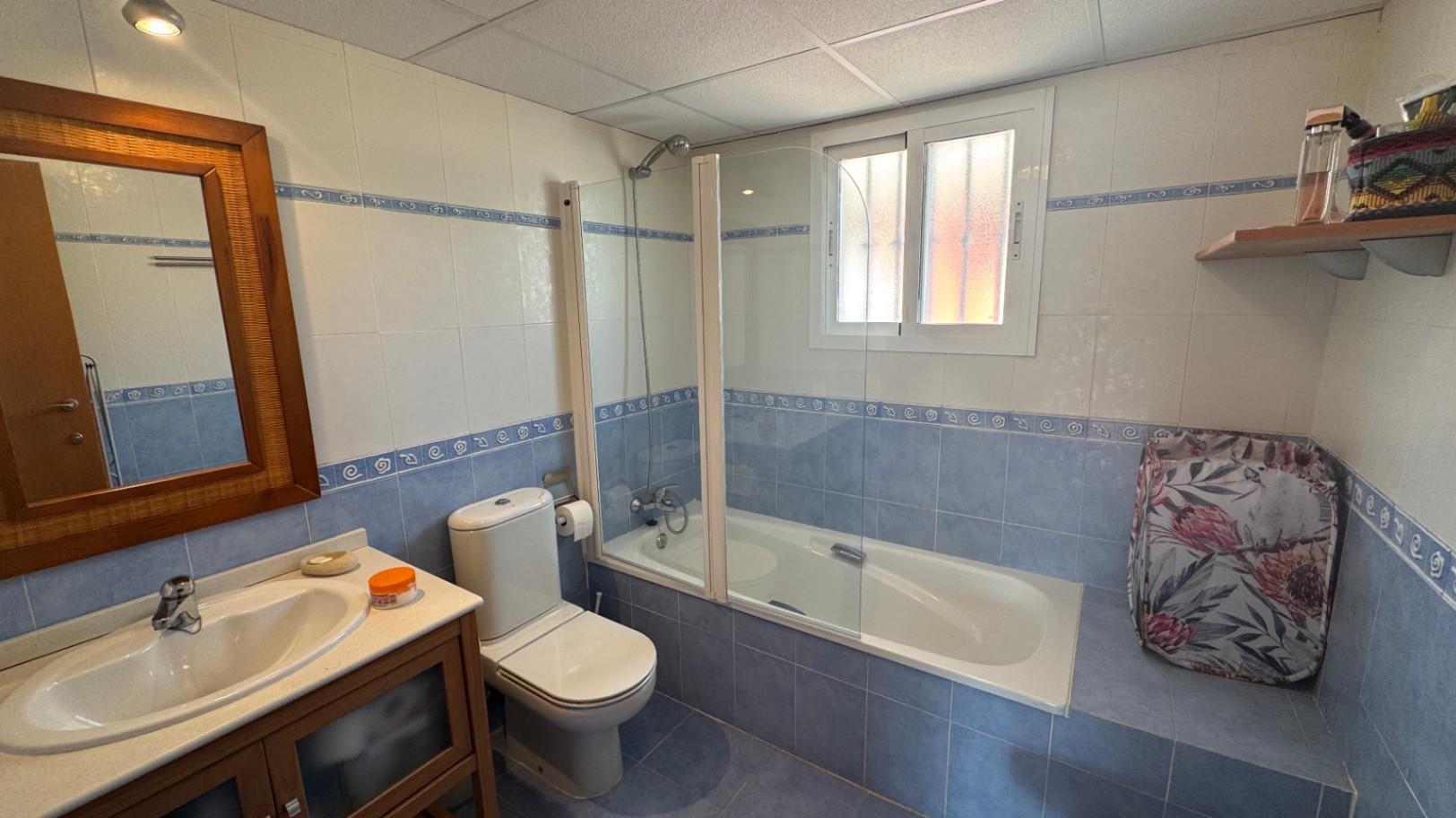 Bathroom featuring blue and white tiling with decorative borders, a bathtub with a glass screen, a wooden-framed mirror, and a matching vanity unit.