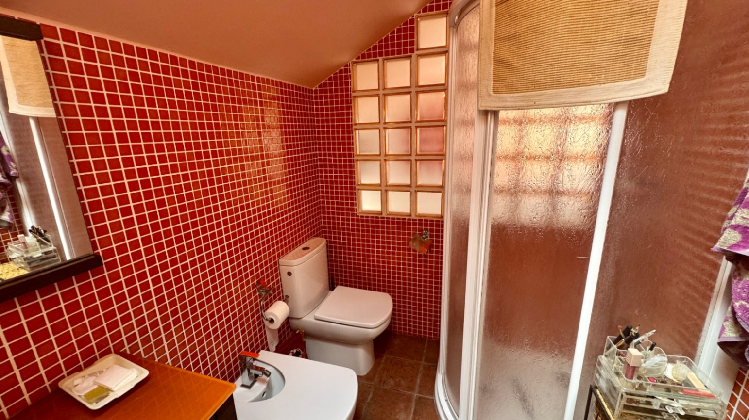 Bathroom featuring red mosaic tile walls, a sloped ceiling, and a curved corner shower. It includes a white toilet, bidet, and a glass block window for diffused light.