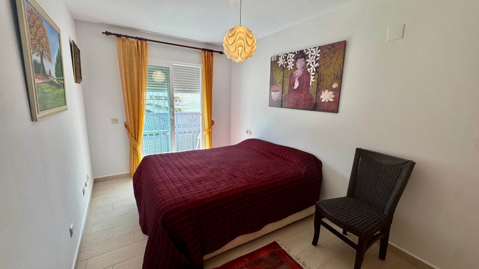 Bedroom with a double bed, red textured bedding, and a dark wicker chair. A sliding glass door with yellow curtains provides natural light and balcony access.
