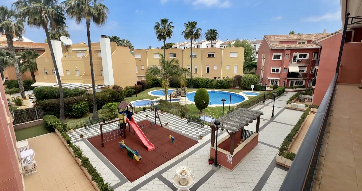 Elevated view of communal grounds featuring a multi-lobed swimming pool, a children's playground with rubber flooring, and landscaped gardens with palm trees.