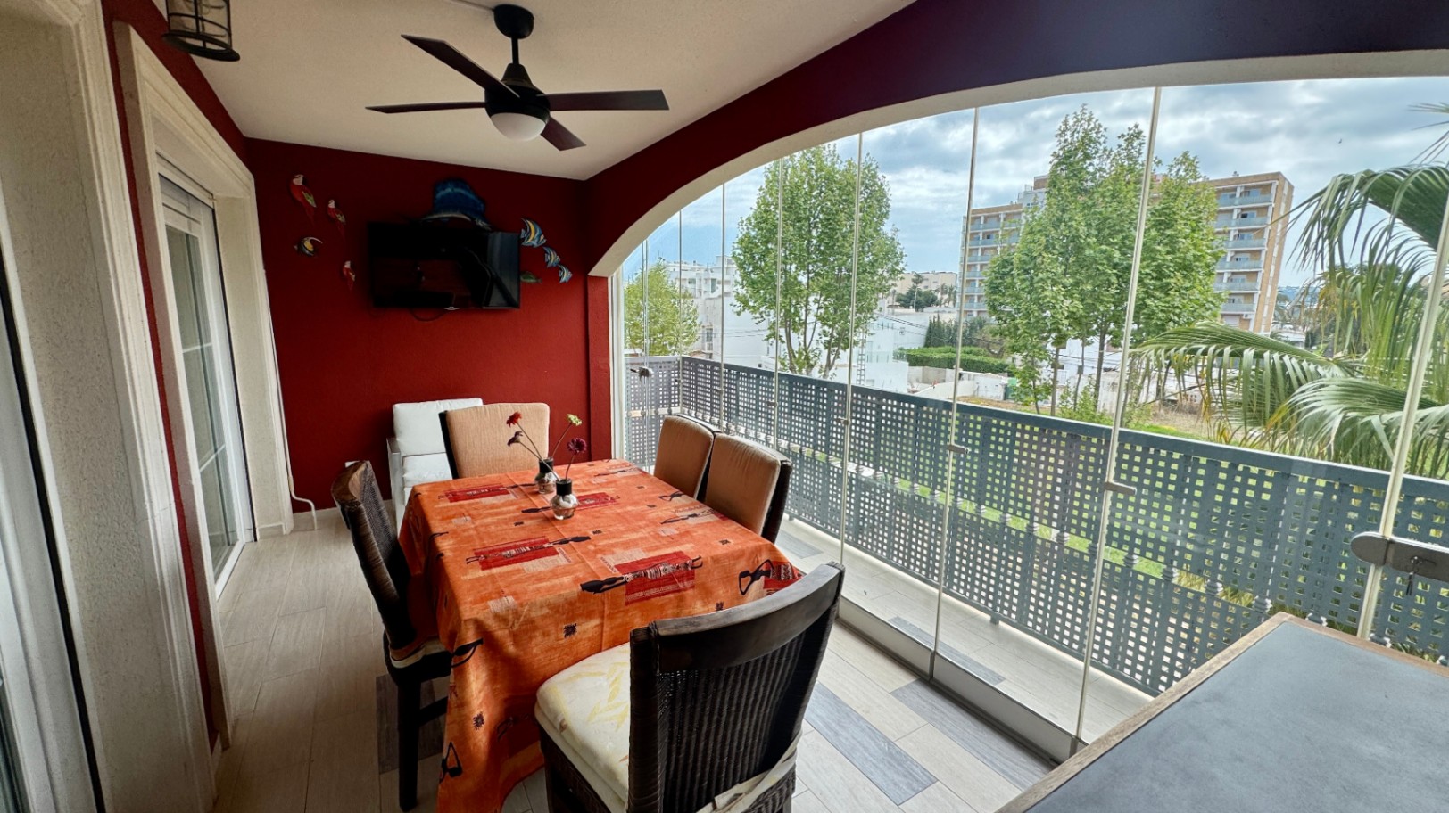 Enclosed terrace featuring glass curtains, a red accent wall with integrated TV, and a dining area overlooking urban greenery and trees.