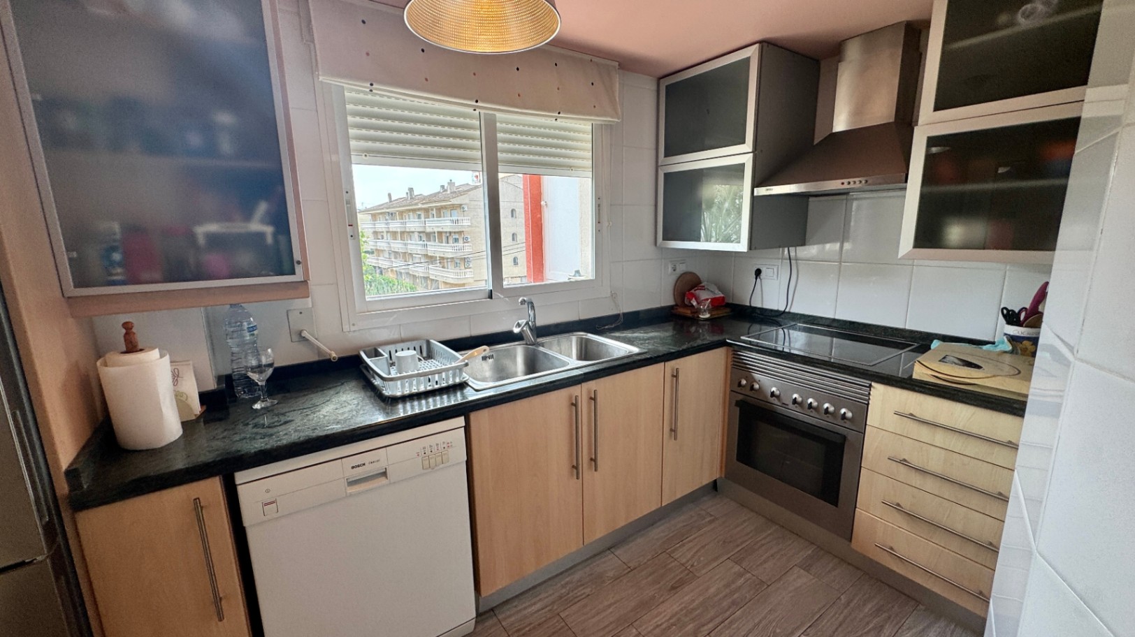 Kitchen featuring light wood cabinetry, dark granite countertops, a Bosch dishwasher, and a stainless steel double sink under a window with urban views.