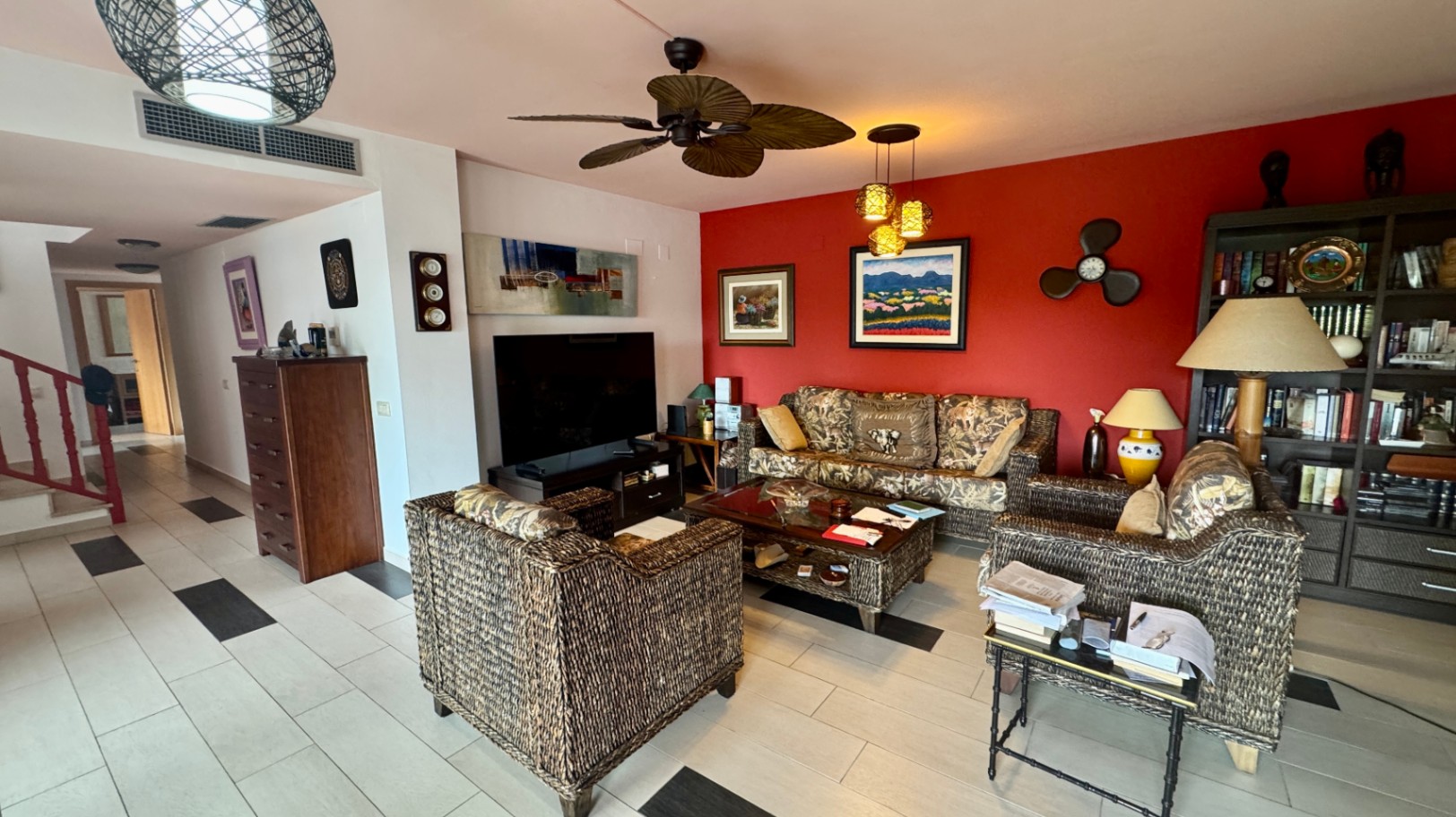 Living room featuring wicker furniture, a bold red accent wall with artwork, and a tropical-style ceiling fan. Light tiled flooring with dark accents connects to the hallway.