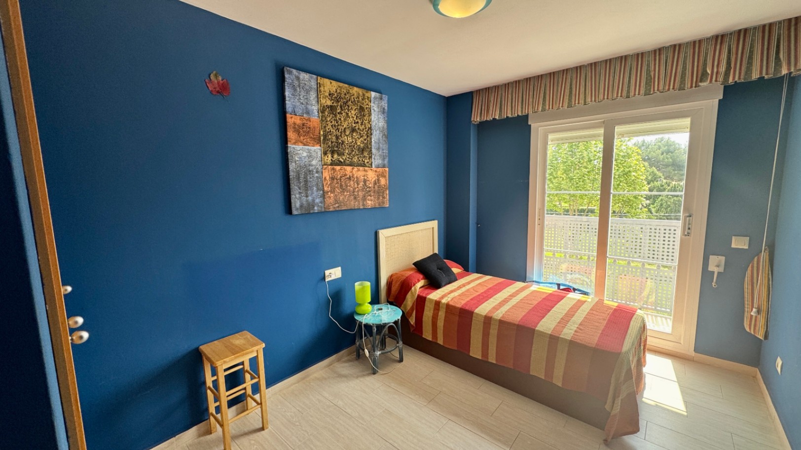 Single bedroom featuring deep blue walls, a bed with a wicker headboard, and a large sliding door opening onto a private balcony with garden views.