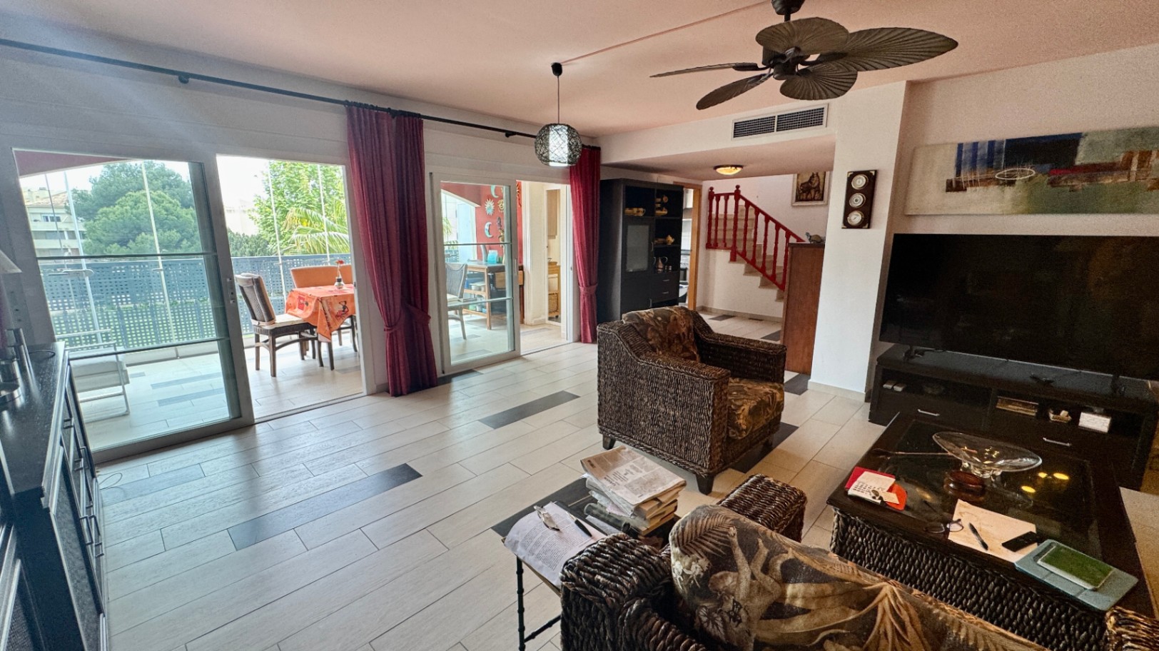 Spacious living area featuring wicker armchairs, tiled flooring with dark accents, and large sliding doors opening onto a furnished terrace. A ceiling fan and red staircase are visible.